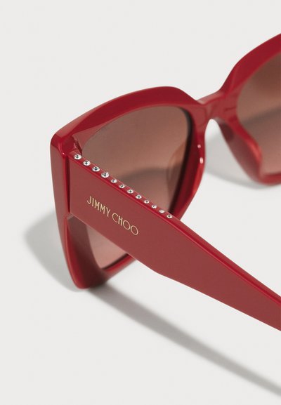 Punased päikeseprillid läikiva viimistlusega, pruuni objektiiviga ja nurga kujuga. Ringi peal on rida sädelevate kividega ning "JIMMY CHOO" bränding kuldse värviga.