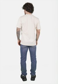 T-shirt a maniche corte color crema con taglio dritto, abbinato a jeans blu e scarpe da ginnastica nere, visto da dietro.