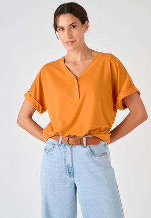 Donna con top arancione a maniche corte con bottoni infilato in jeans azzurri con cintura marrone, in posa con le mani sui fianchi.