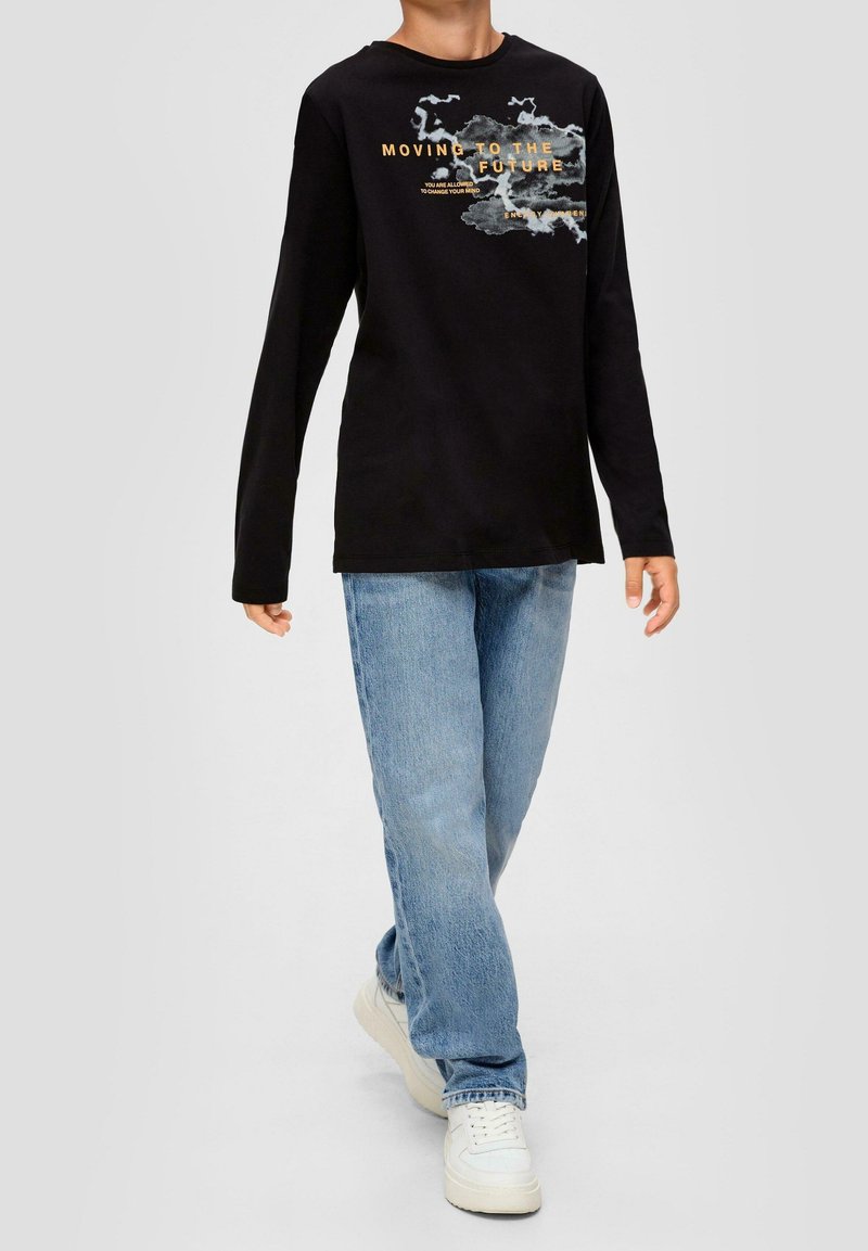 Enfant portant un t-shirt noir à manches longues avec le texte « MOVING TO THE FUTURE » et un visuel, un jean bleu clair et des baskets blanches, marchant vers l'avant.
