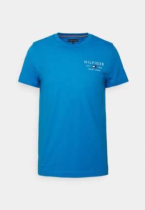 Niebieski t-shirt z krótkim rękawem i okrągłym dekoltem, biały tekst "Hilfiger Est. 1985 New York" oraz małe logo flagi na górnej lewej stronie klatki piersiowej.