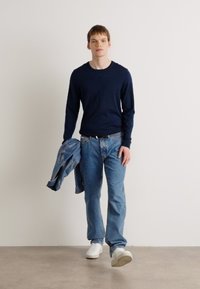 Cămașă albastră cu mânecă lungă, asortată cu blugi drepti de culoare albastru deschis și teniși albi, ținând o jachetă din denim. Design simplu și casual.