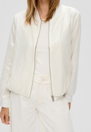 Vrouw draagt een wit gestructureerd jack met rits over een witte geribbelde top en witte broek met hoge taille, handen ontspannen langs het lichaam.