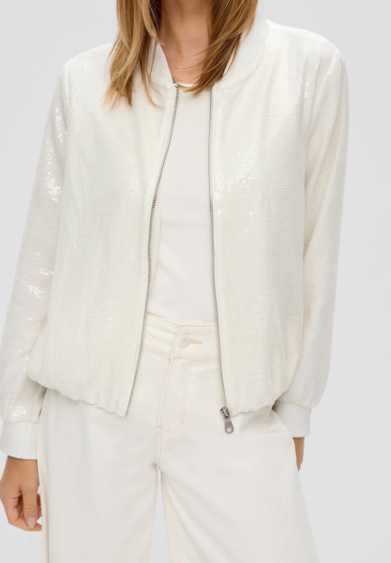 Femme portant une veste zippée blanche texturée par-dessus un haut côtelé blanc et un pantalon blanc taille haute, les mains détendues le long du corps.