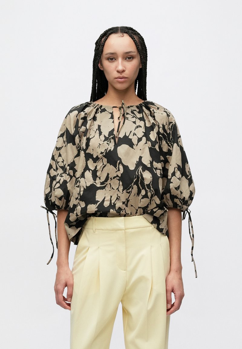 Luźna, czarno-beżowa bluzka w kwiatowy wzór, z dekoltem w szpic i troczkami, zestawiona z jasnożółtymi spodniami.