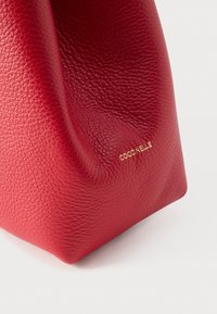 Punane nahast kott, mille pinnal on tekstuur ja pehmed voldid; esiküljel on kuldse logoga reljeef, kott seisab lamedal alusel.