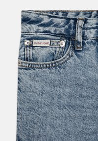 Svijetloplave traperice s teksturiranim završetkom, s crvenom etiketom "Calvin Klein", metalnim gumbima i prednjim džepom.