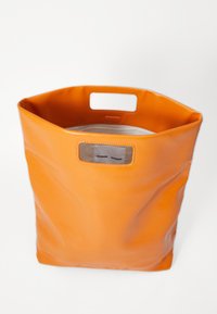 Oranžna usnjena torba v obliki heksagona, ki ima šivano šivanko in oznako logotipa znotraj. Ima ročaje na vrhu za nošenje.