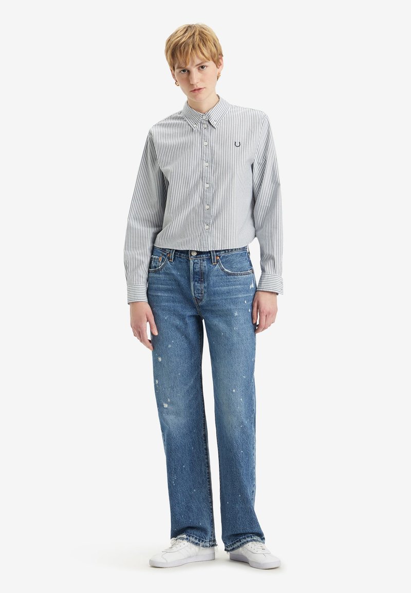 Camicia a righe button-down azzurro chiaro e bianca, abbinata a jeans a gamba larga azzurro chiaro strappati e sneakers bianche. Outfit casual.