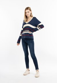 Pulover în dungi navy cu accente crem și roșii, design cu decolteu în V, manșete canelate, purtat cu blugi închiși și adidași deschiși. Textură moale.