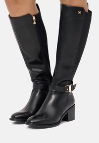 Botas negras hasta la rodilla, hechas de cuero suave, con una cremallera lateral, acentos de hardware dorado y una correa texturizada en el tobillo. Tacón cuadrado.