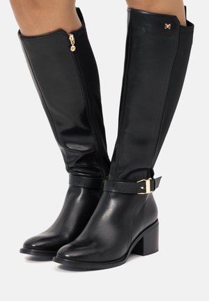 Botas negras hasta la rodilla, hechas de cuero suave, con una cremallera lateral, acentos de hardware dorado y una correa texturizada en el tobillo. Tacón cuadrado.
