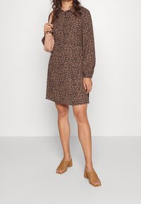 Brun blommig klänning med krage och långa ärmar, kombinerad med sandfärgade vävda tofflor och en ljusbeige handväska.