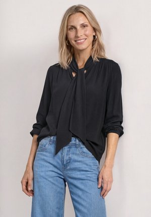 Femme souriante aux cheveux blonds mi-longs, portant un chemisier noir avec une cravate et un jean bleu clair, debout devant un fond clair uni.