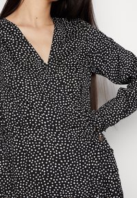 Svart omlotklänning med vita prickar, långa ärmar, V-ringning och knytning i midjan. Tillverkad av ett lätt tyg med en slät struktur.