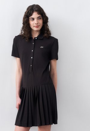Femme aux cheveux foncés bouclés portant une robe polo noire à manches courtes avec des boutons et une jupe plissée, debout contre un mur blanc.