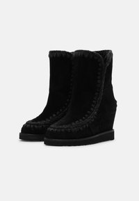 Botines de tacón cuña negros hechos de ante. Presentan un interior acolchado y un pespunte decorativo a lo largo del borde. La suela de goma plana proporciona estabilidad.