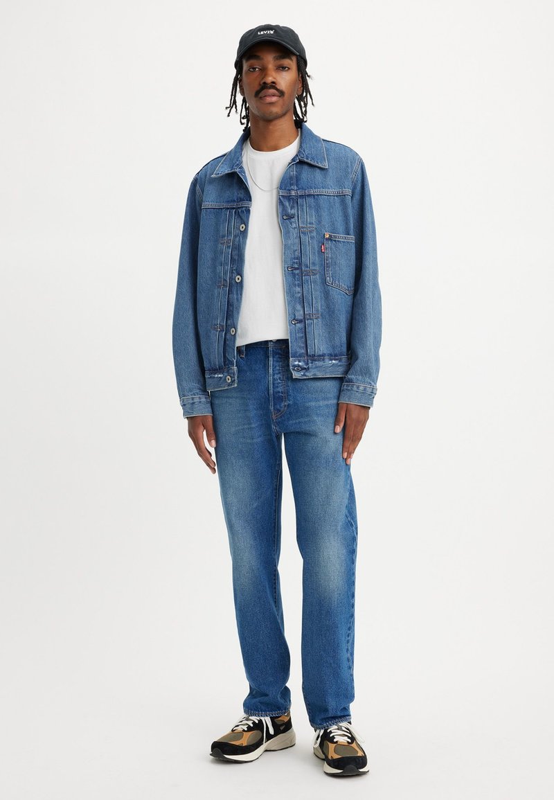 Veste en denim bleu avec un col et une poche avant, sur un t-shirt blanc, assortie à un jeans bleu clair et des baskets noir et blanc. Casquette noire.
