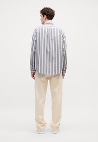 Cămașă cu dungi, gri și alb, cu un hem curbat, asortată cu pantaloni largi de culoare crem și adidași albi. Vedere din spate.