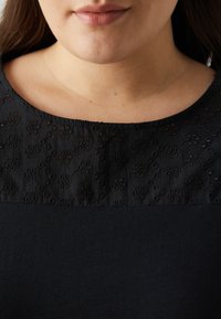 Černý top s texturovanou květinovou výšivkou podél výstřihu, zatímco zbytek oděvu je pokryt hladkým materiálem.
