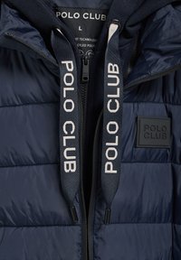 Mörkblå vadderad jacka med dragkedja, logotypdrapeade snören och ett litet märkt märke på framsidan. Tillverkad av återvunnen polyester.