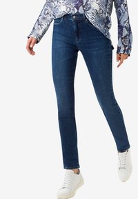 Jeans skinny de mezclilla azul oscuro con tiro medio, cinco bolsillos y un ligero estiramiento. Combinados con una blusa estampada y zapatillas blancas.