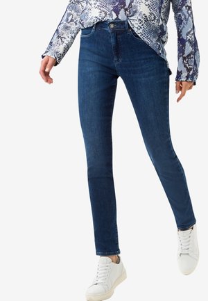 Jean skinny en denim bleu foncé avec une taille moyenne, cinq poches et une légère élasticité. Porté avec un haut à motifs et des baskets blanches.