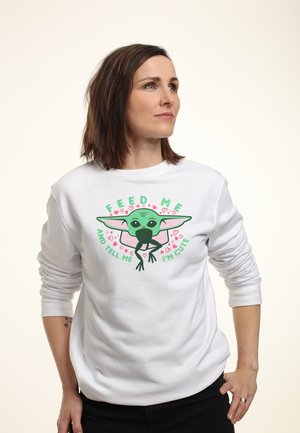 Sudadera blanca con un personaje de dibujos animados que tiene orejas grandes y piel verde, rodeado del texto "ALIMÉNTAME Y DIME QUE SOY LINDO" en verde y rosa.