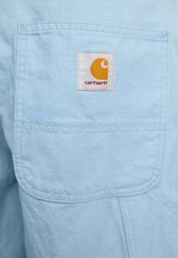 Lichtblauwe stof met een textuurafwerking, voorzien van een wit label met een oranje logo en rode tekst in het midden. Zichtbare stiksels.