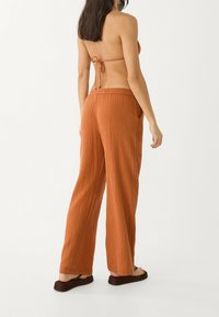 Pantalon ample de couleur rouille, texturé, avec une taille à cordon et des poches latérales, porté avec des sandales plates. Vue de dos affichée.