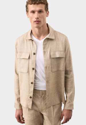 Mann trägt beigen Hemdjacke mit Knöpfen und Fronttaschen über weißem V-Ausschnitt-T-Shirt und passende beige Hose, steht vor einfachem Hintergrund.
