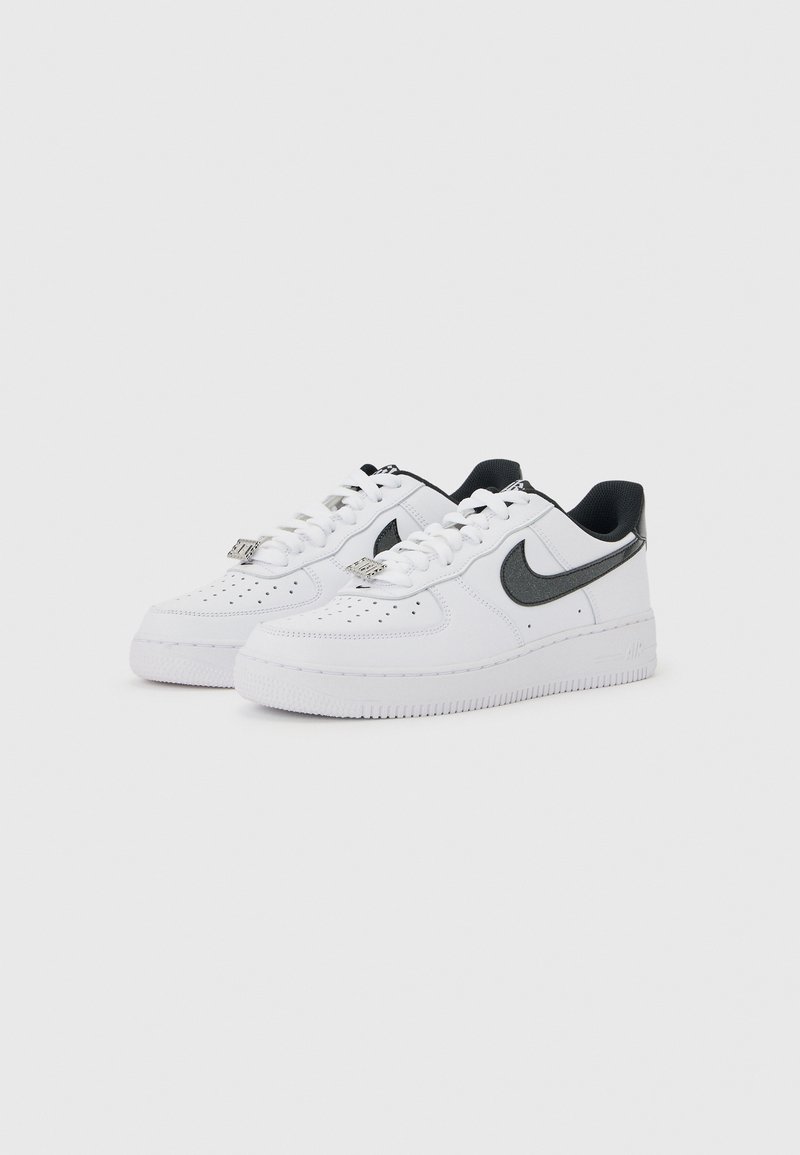 Paire de baskets basses blanches avec logos Nike swoosh noirs et lacets blancs sur fond gris clair uni.