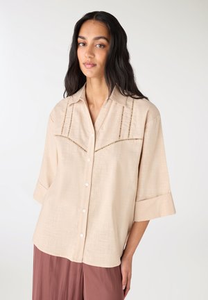 Donna con capelli lunghi e scuri indossa una camicia beige con bottoni e dettagli ritagliati e pantaloni larghi color malva, in piedi davanti a uno sfondo chiaro e uniforme.