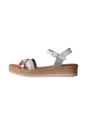 Sandalia cuña con tiras blancas y metálicas, hebilla ajustable en el tobillo, plantilla color cuero y suela intermedia de yute trenzado sobre fondo blanco.