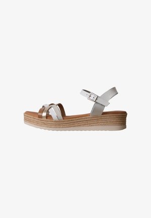 Sandalia cuña con tiras blancas y metálicas, hebilla ajustable en el tobillo, plantilla color cuero y suela intermedia de yute trenzado sobre fondo blanco.