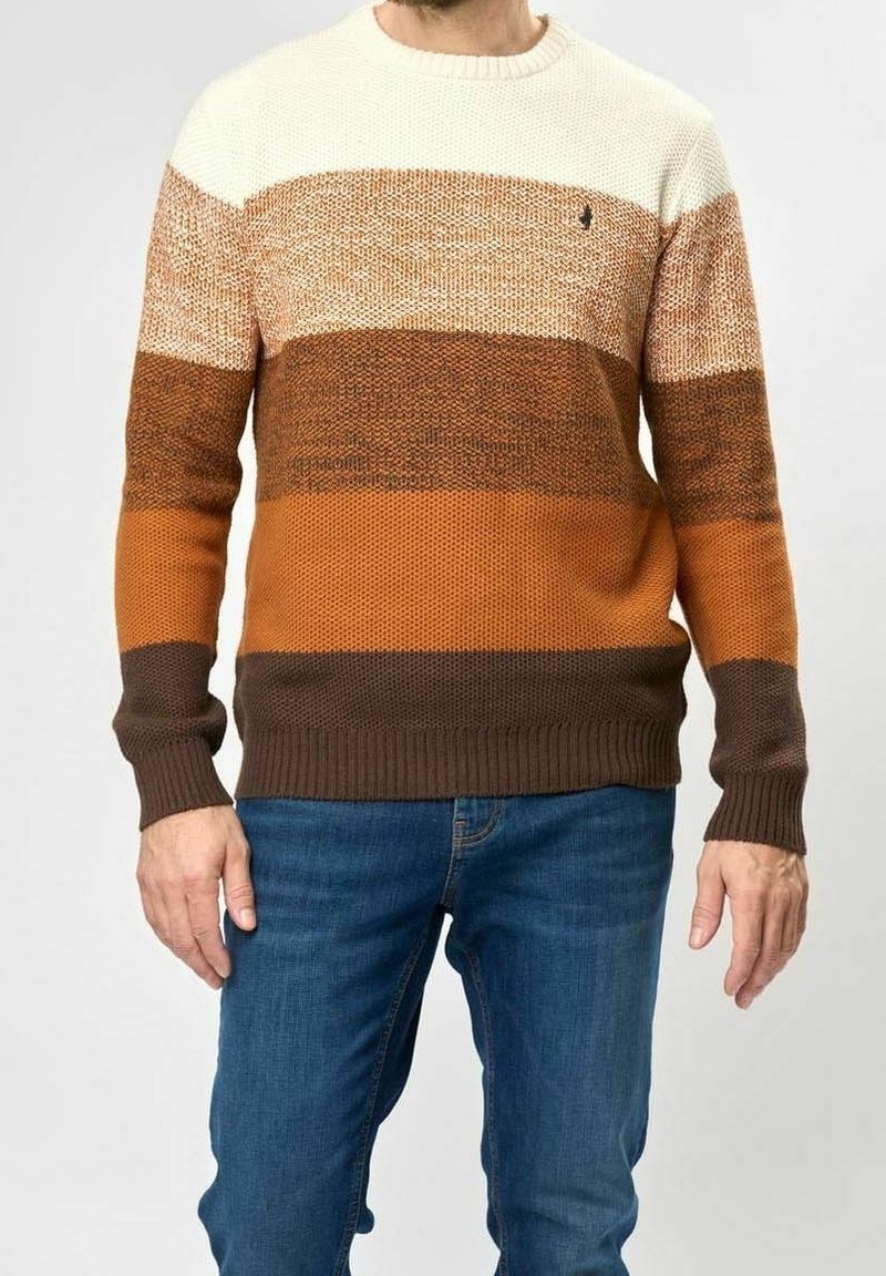 Homme portant un pull rayé marron, orange, beige et crème avec un jean bleu, debout devant un fond uni.