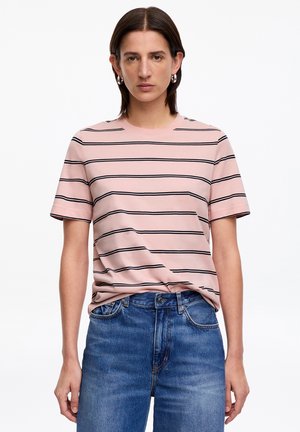 Kvinde iført en lyserød kortærmet skjorte med sorte vandrette striber og blå denimjeans, stående mod en hvid baggrund.