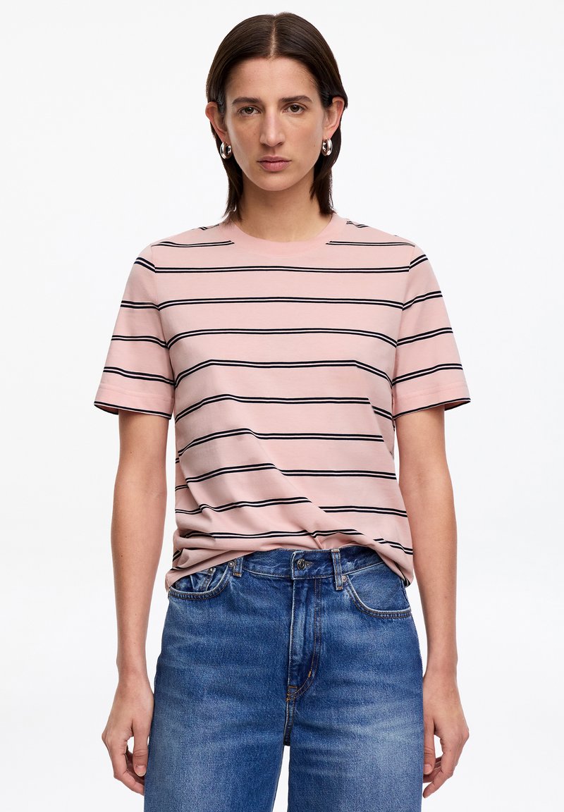 Femme portant un t-shirt rose à manches courtes avec des rayures horizontales noires et un jean en denim bleu, debout devant un fond blanc.