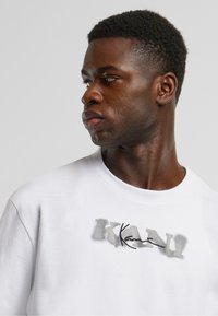 White cotton t-shirt with a crew neckline, featuring a grey graphic print of "KANI" across the chest. Smooth texture, relaxed fit.