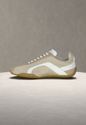 Sneaker en cuir beige et blanc avec des lacets blancs et une semelle gomme, présenté de profil sur une surface claire avec un arrière-plan neutre.