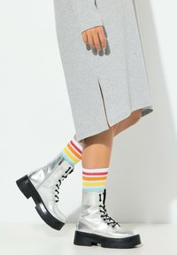 Hopeanväriset metallinhohtoiset nauhalliset saappaat mustilla paksuilla pohjilla, yhdistettynä monivärisiin raidallisiin sukkiin, joissa on punaisen, keltaisen ja sinisen sävyt, harmaan mekon alla.