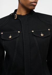 Κοντινή λήψη ενός ατόμου που φοράει μαύρο σακάκι με χρυσά κουμπιά, τσέπες με πτερύγια, αντίθετη λευκή ραφή και χρυσό κούμπωμα στον γιακά.