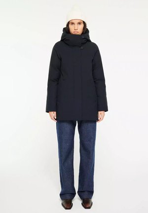 Zwarte pufferjack met een hoge kraag, voorzien van twee knopen, gecombineerd met brede blauwe spijkerbroeken en een crèmekleurige gebreide beanie.