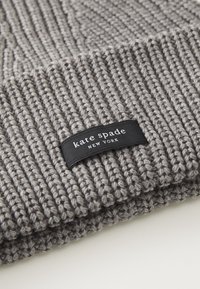 Šedá pletená čiapka s rebrovanou textúrou a čiernou látkovou nášivkou s nápisom "kate spade NEW YORK". Obsahuje hrubú, silnú priadzu.