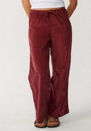 Persoon draagt loszittende bordeaux corduroy broek en open teen bruine sandalen en staat met handen achter de rug.