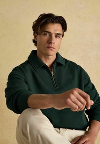 Zielony pulower z zamkiem na szyi, wykonany z miękkiego materiału, z kołnierzykiem i ściągaczami na rękawach, zestawiony z jasnymi spodniami na neutralnym tle.