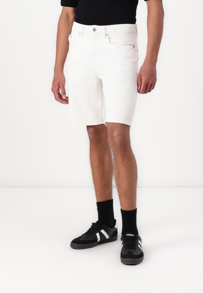 White denim shorts with a straight cut and frayed hem, featuring a waist button, five pockets, and styled with black footwear.
