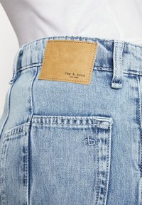 Los jeans de mezclilla azul claro presentan un parche de cuero marrón claro con el logotipo "rag & bone" en relieve. La tela tiene una textura suave y un diseño clásico de cinco bolsillos.