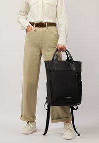 Czarny plecak-torba z wyściełanymi paskami, górnymi uchwytami, przednią kieszenią na zamek i gładką fakturą. Noszony przez osobę w jasnych spodniach i białej koszuli.