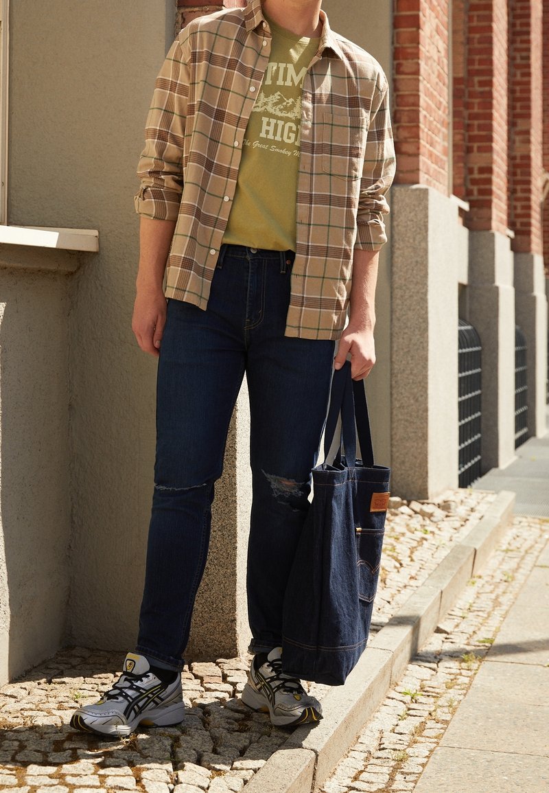 Tenue décontractée comprenant une chemise à carreaux beige portée sur un t-shirt graphique vert, un jean bleu foncé avec des effets usés, et des baskets grises. Tenant un sac fourre-tout en denim.